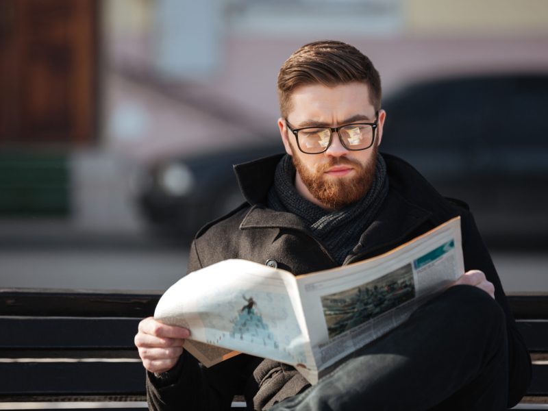 Concentrated bearded young man in glasses sitting on bench and reading newspaper outdoors